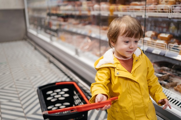 Bambino al mercato con un carrello della spesa che indossa una giacca gialla e jeans