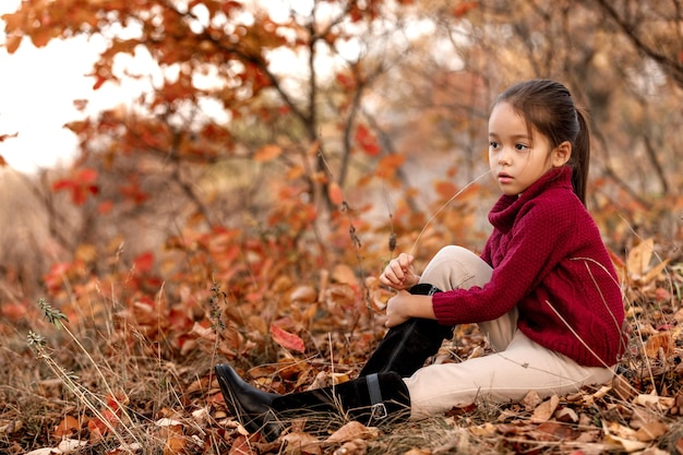 Bambino alla moda di 5 anni in posa nel parco autunnale. bambina seduta tra le foglie cadute