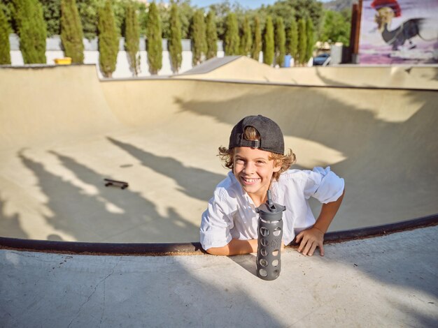 Bambino allegro con una bottiglia d'acqua che guarda l'obbiettivo e appoggiato sulla rampa nello skate park in una giornata di sole