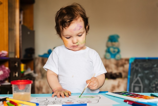 Bambino allegro felice che disegna con il pennello nell'album usando molti strumenti di pittura.