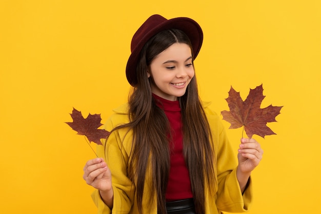 Bambino allegro in cappello e cappotto con foglie d'acero autunnali su sfondo giallo, caduta.