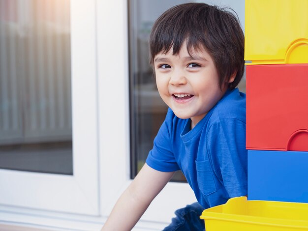 Bambino attivo del ritratto che si nasconde accanto alla scatola di plastica variopinta che gioca nascondino, bambino felice divertendosi giocando nella stanza del gioco. Ragazzo di 6 anni che si rilassa a casa nel fine settimana. Bambini positivi