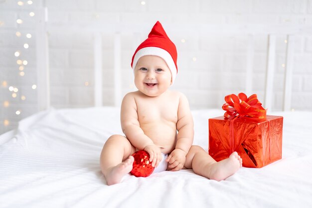 bambino bambino in rosso santa berretto si siede con un regalo su uno sfondo bianco capodanno vacanze di natale