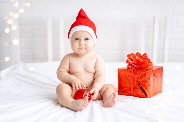 bambino bambino in rosso santa berretto si siede con un regalo su uno sfondo bianco capodanno vacanze di natale