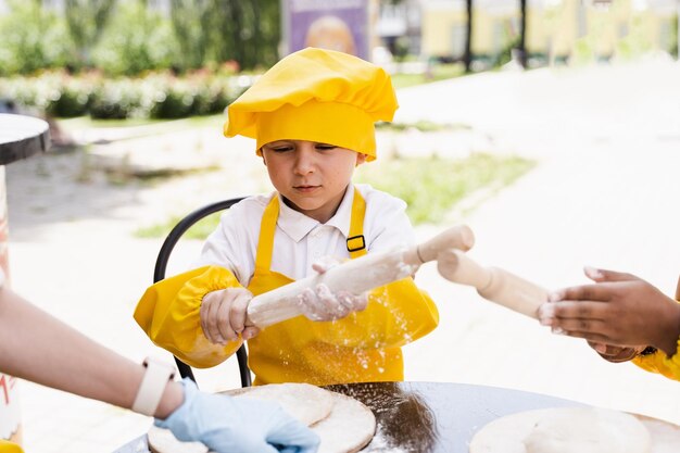 Bambino bello del cuoco in cappello giallo del cuoco unico e uniforme gialla del grembiule che cucina e che tiene il rullo della pasta all'aperto