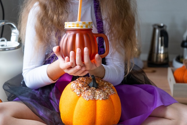 Bambino beve succo di zucca frullato di verdure o cocktail da una tazza con una cannuccia La cucina decora per Halloween Ragazza in un costume da strega Autunno accogliente in casa