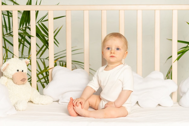 Bambino biondo con gli occhi azzurri in un body bianco su un letto con biancheria intima di cotone a casa prima di andare a letto