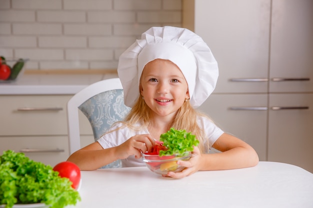 bambino biondo in un cappello da chef in cucina a mangiare verdure