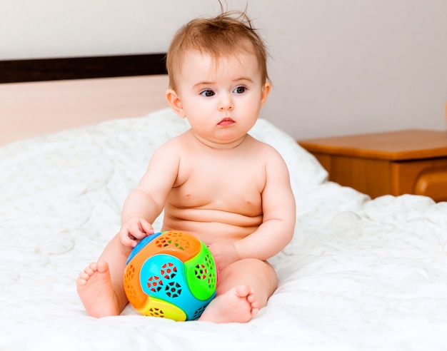 Bambino biondo sveglio che gioca con una palla seduto sul letto in camera da letto. bambino felice di 6 mesi che gioca con una palla