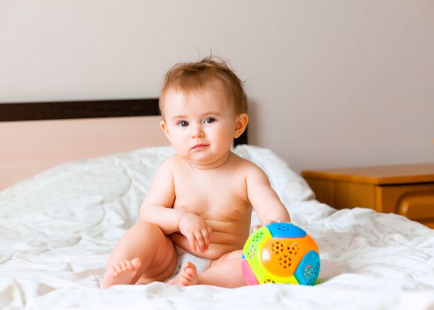 Bambino biondo sveglio che gioca con una palla seduto sul letto in camera da letto. bambino felice di 6 mesi che gioca con una palla