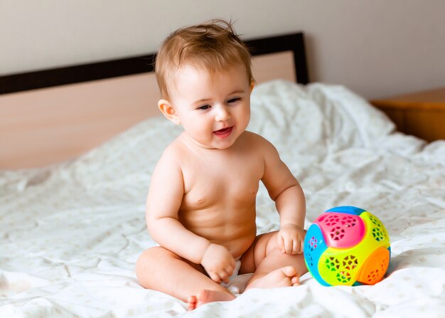Bambino biondo sveglio che gioca con una palla seduto sul letto in camera da letto. bambino felice di 6 mesi che gioca con una palla