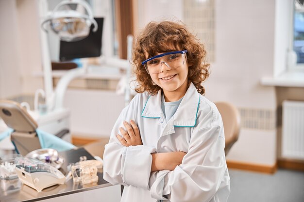 Bambino bruna riccio che indossa un camice da laboratorio e occhiali di sicurezza e in piedi con le braccia incrociate nell'ufficio del dentista