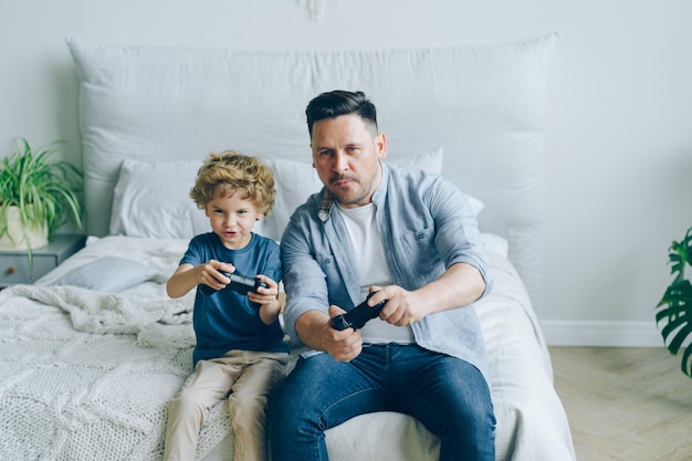 Bambino carino che gioca al videogioco con il padre che tiene i joystick divertendosi a letto