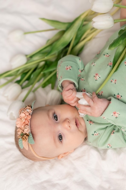 Bambino carino con una fascia floreale e un body di cotone verde a casa su un letto bianco con tulipani. spr