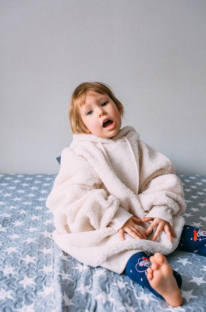 Bambino carino y sul letto in comodi vestiti per adulti e un cappello con paraorecchie. Emozioni e stile di vita domestico