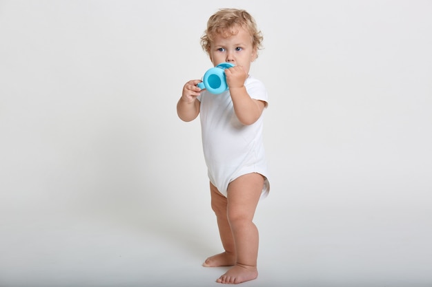 Bambino che beve acqua dal biberon blu, bambino che indossa una tuta bianca, distoglie lo sguardo, in piedi a piedi nudi contro il muro luminoso, affascinante bambino neonato ha sete.