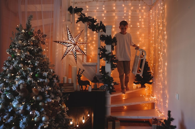 Bambino che decora l'albero di Natale a casa. Ragazzino in maglione lavorato a maglia con ornamento di Natale. La famiglia con bambini celebra le vacanze invernali. I bambini decorano il soggiorno e il camino per Natale.