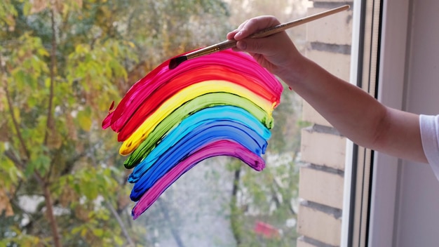 Bambino che dipinge a mano arcobaleno colorato sulla finestra durante la quarantena Covid19 a casa