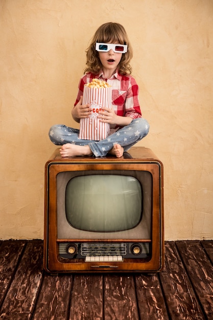 Bambino che gioca in casa. Bambino seduto sulla TV retrò e mangiare popcorn. Concetto di cinema