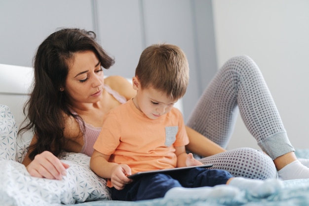 Bambino che guarda i cartoni animati su tablet mentre era seduto sul letto mentre sua madre guardava i cartoni animati con lui. Interno camera da letto.