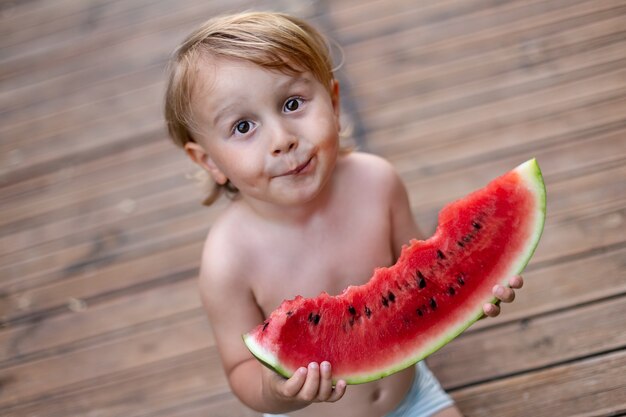 Bambino che mangia anguria in giardino durante le vacanze estive. I bambini mangiano frutta all'aperto. Spuntino sano per i bambini. Ragazzino che assaggia una fetta di anguria.