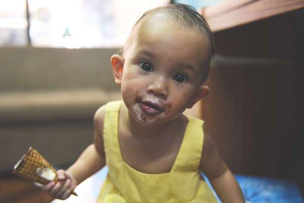 Bambino che mangia il gelato / cialda felice del cono gelato della tenuta della neonata asiatica divertente con il viso e la bocca sporchi