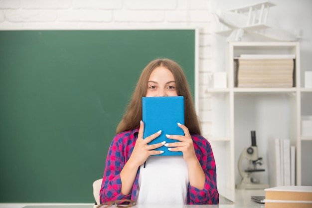 Bambino che si nasconde dietro il libro a scuola sulla conoscenza di base della lavagna