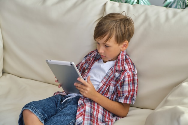 Bambino che tiene e gioca sul suo dispositivo tablet seduto sul divano bianco, godendosi il tempo libero, in soggiorno, indossando jeans e una camicia a quadri