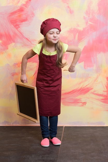 Bambino chef in grembiule su sfondo colorato. Cuoco di piccola ragazza in cappello con lavagna e cucchiaio di legno. Ristorante e menu design, copia spazio. Cucina ed educazione. Felicità e infanzia.