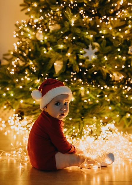 Bambino con cappello da Babbo Natale, seduto su un albero di Natale con sfondo di luci, a casa, al buio, che guarda l'obbiettivo.