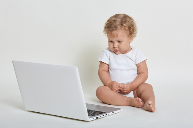 Bambino con computer portatile seduto sul pavimento isolato sopra il muro bianco