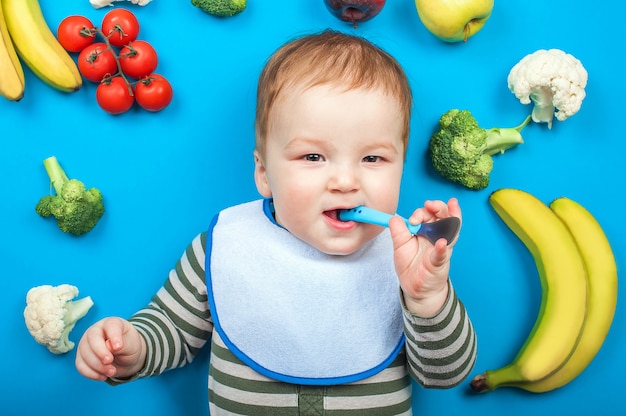 Bambino con frutta e verdura su sfondo blu, alimenti per bambini sani. Omogeneizzato, banane, broccoli, cavolfiori, zucchine, mele, pomodori