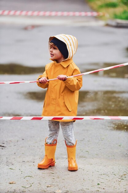 Bambino con mantello impermeabile giallo e stivali che giocano all'aperto vicino a nastro protettivo dopo la pioggia