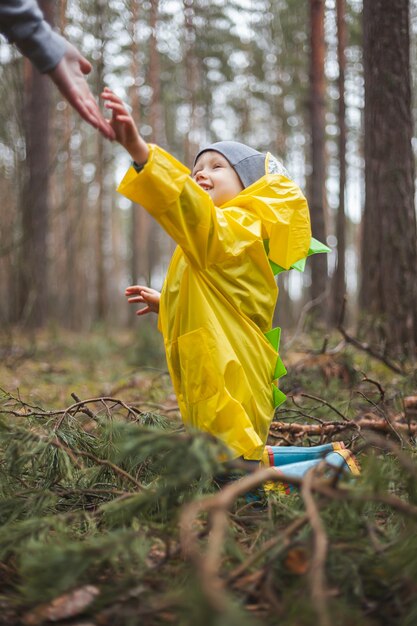 Bambino con un impermeabile giallo che cammina nella foresta dopo la pioggia, si siede a terra tra i rami di pino, la mamma lo raggiunge e lo aiuta ad alzarsi