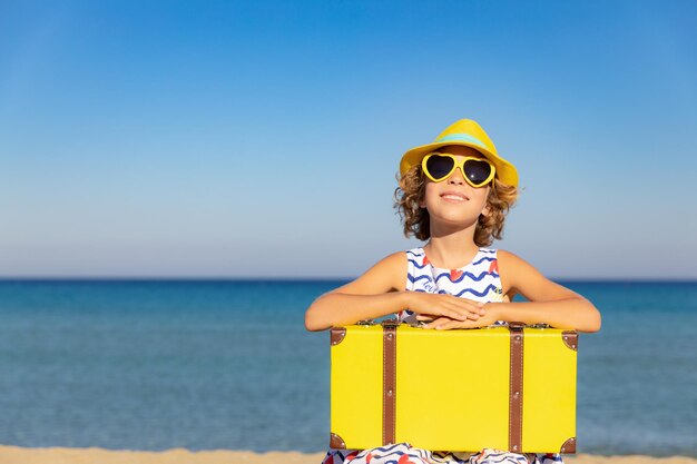 Bambino con valigia vintage in vacanza estiva Ragazza che si diverte sulla spiaggia Bambino seduto sullo sfondo del mare e del cielo Concetto di viaggio e avventura