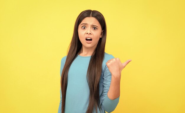 Bambino confuso con i capelli lunghi punta il dito sullo spazio della copia su sfondo giallo emozioni