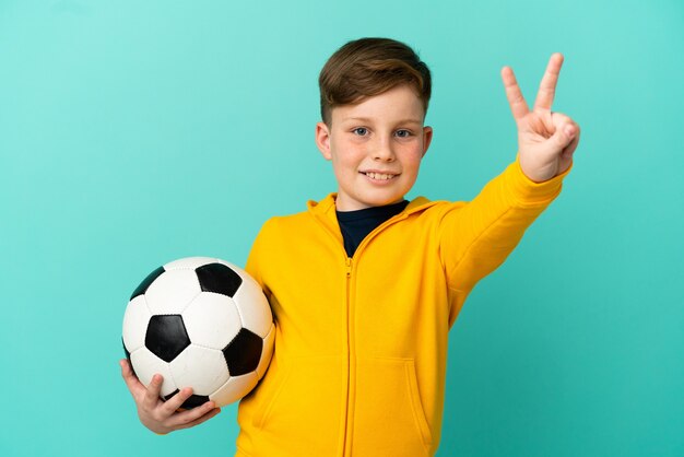 Bambino dai capelli rossi che gioca a calcio isolato sulla superficie blu che sorride e che mostra il segno della vittoria