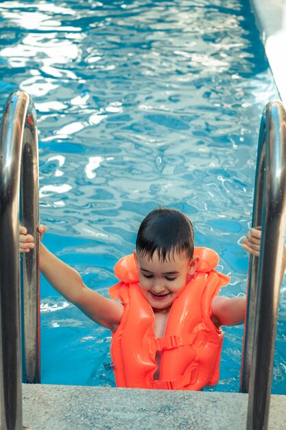 Bambino delizioso felice con i capelli bagnati in piedi sul corrimano cromato in acqua dolce nuoto giubbotto di salvataggio all'aperto piscina arancione nella giornata di svago di vacanza estiva. Attività estive per bambini felici in piscina