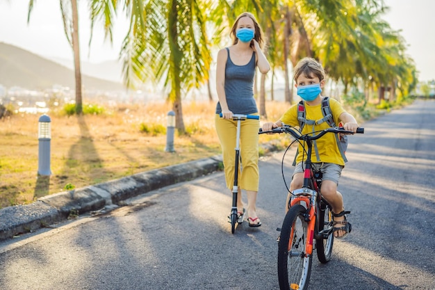 Bambino della scuola attivo e sua madre in maschera medica in sella a una bicicletta con zaino in una giornata di sole Bambino felice in bicicletta sulla strada per la scuola Devi andare a scuola con una maschera a causa dell'epidemia di coronavirus