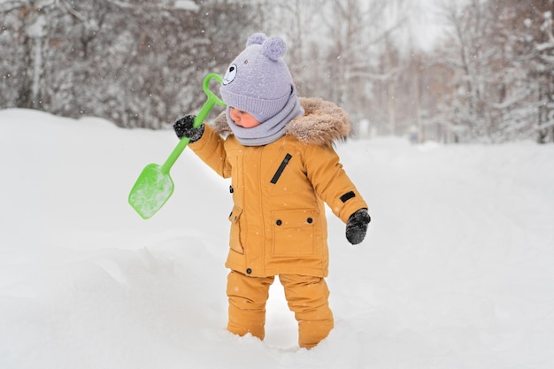 Bambino di 1217 mesi con una pala giocattolo verde nelle sue mani che gioca con la neve in un parco invernale