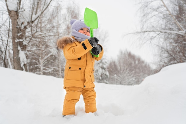 Bambino di 1217 mesi in abiti gialli caldi che tiene in mano una pala da neve in plastica giocattolo