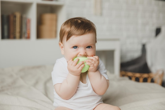 Bambino di 6 mesi rosicchia la palla tenendola con le mani seduto sul letto a denti