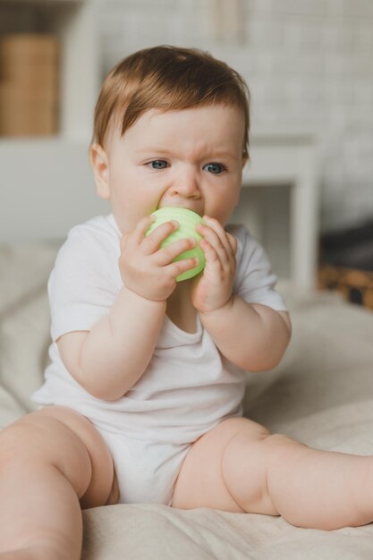 Bambino di 6 mesi rosicchia la palla tenendola con le mani seduto sul letto a denti