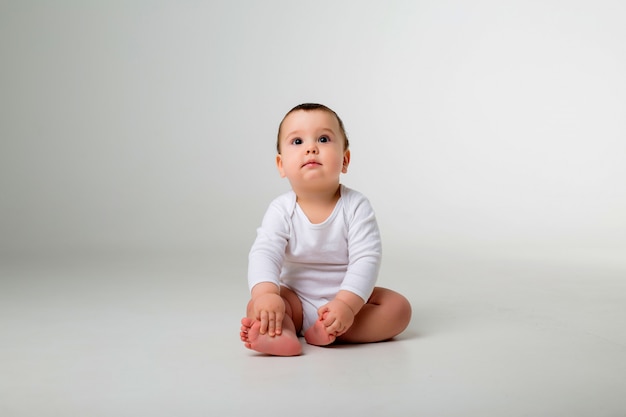 bambino di 9 mesi in un body bianco seduto su un muro bianco