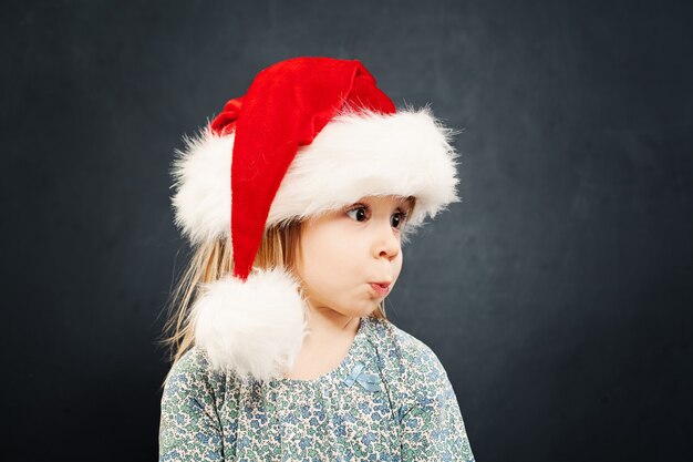 Bambino di Natale sorpreso in cappello da Babbo Natale su sfondo lavagna