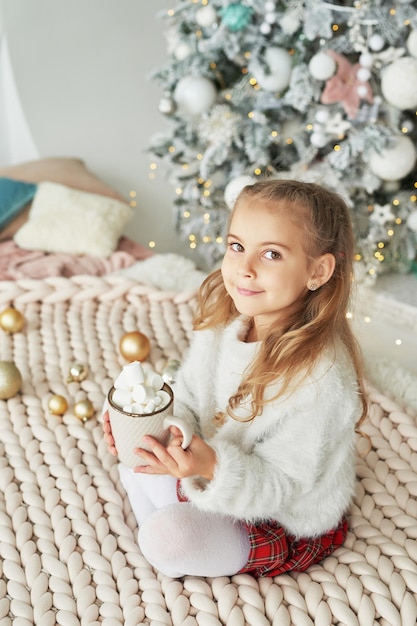 Bambino di Natale sullo sfondo dell'albero di Natale con una tazza di cacao e marshmallow.