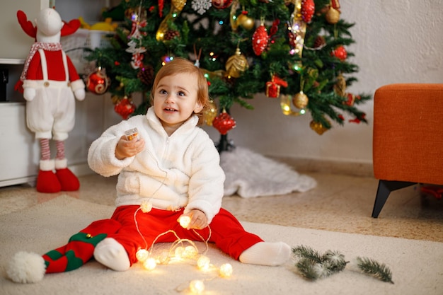 Bambino divertente che gioca con la ghirlanda di Natale su un tappeto morbido davanti a un albero di Natale