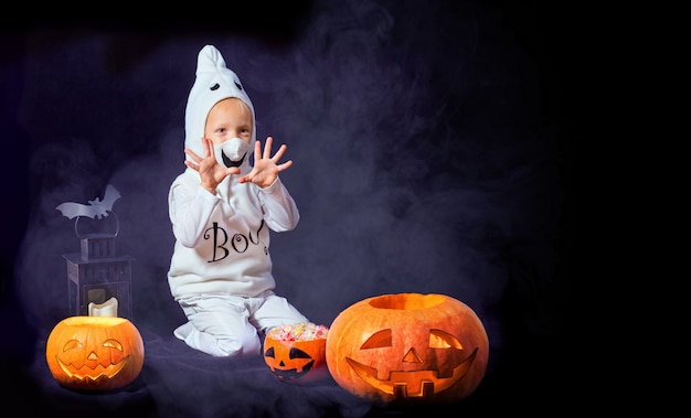 Bambino divertente in un costume di carnevale bianco di un fantasma nella stanza. Ragazzo che gioca con zucche e caramelle.