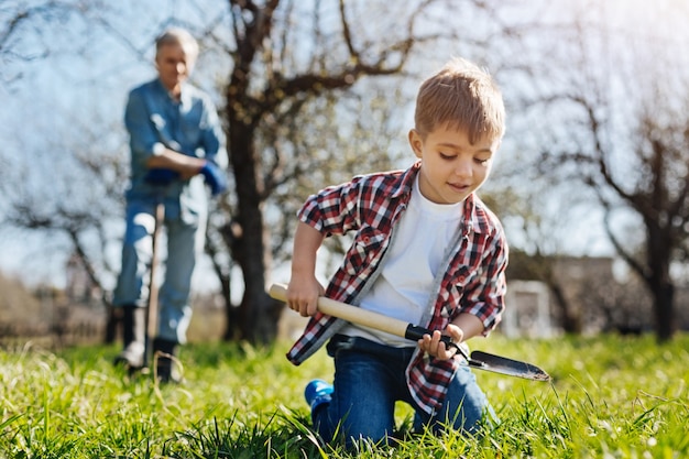 Bambino eccitato che indossa una camicia a quadri rossa in piedi sulle ginocchia e scavare il terreno mentre si diverte con suo nonno in piedi dietro