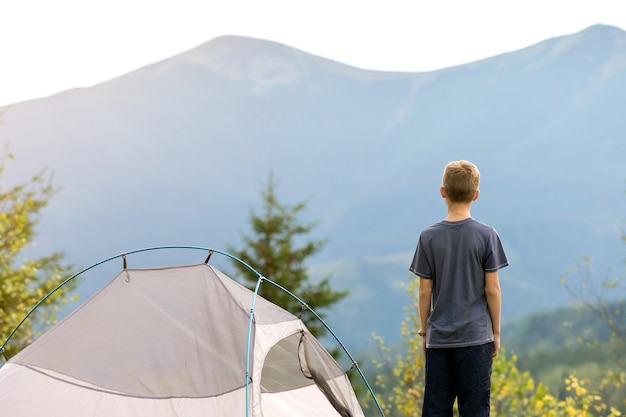 Bambino escursionista che riposa in piedi vicino a una tenda da campeggio in un campeggio di montagna che gode della vista della natura estiva.